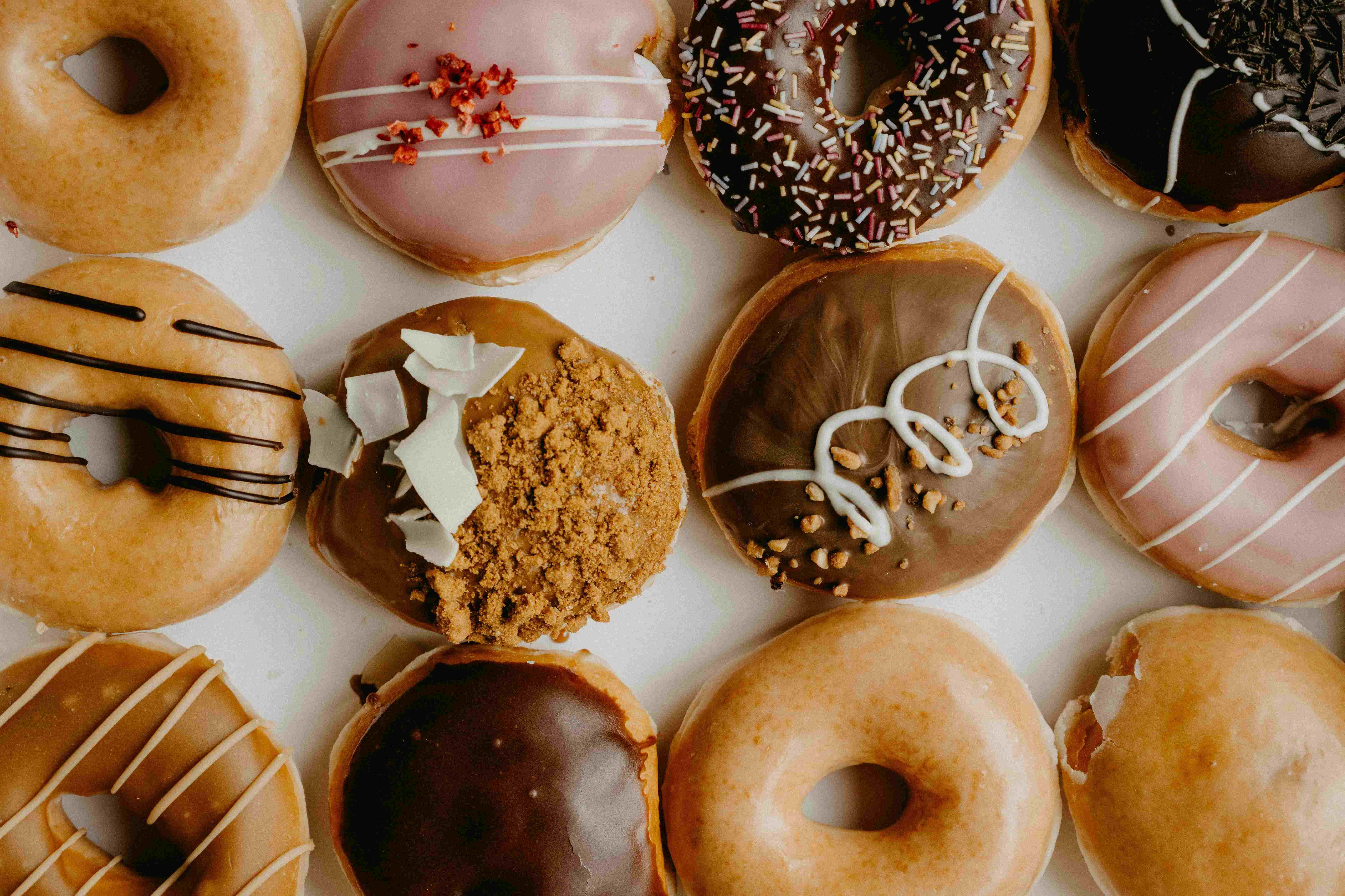Varietà di ciambelle glassate e farcite disposte su sfondo bianco, con diverse decorazioni tra cui cioccolato, granella, zuccherini colorati e glasse rosa.