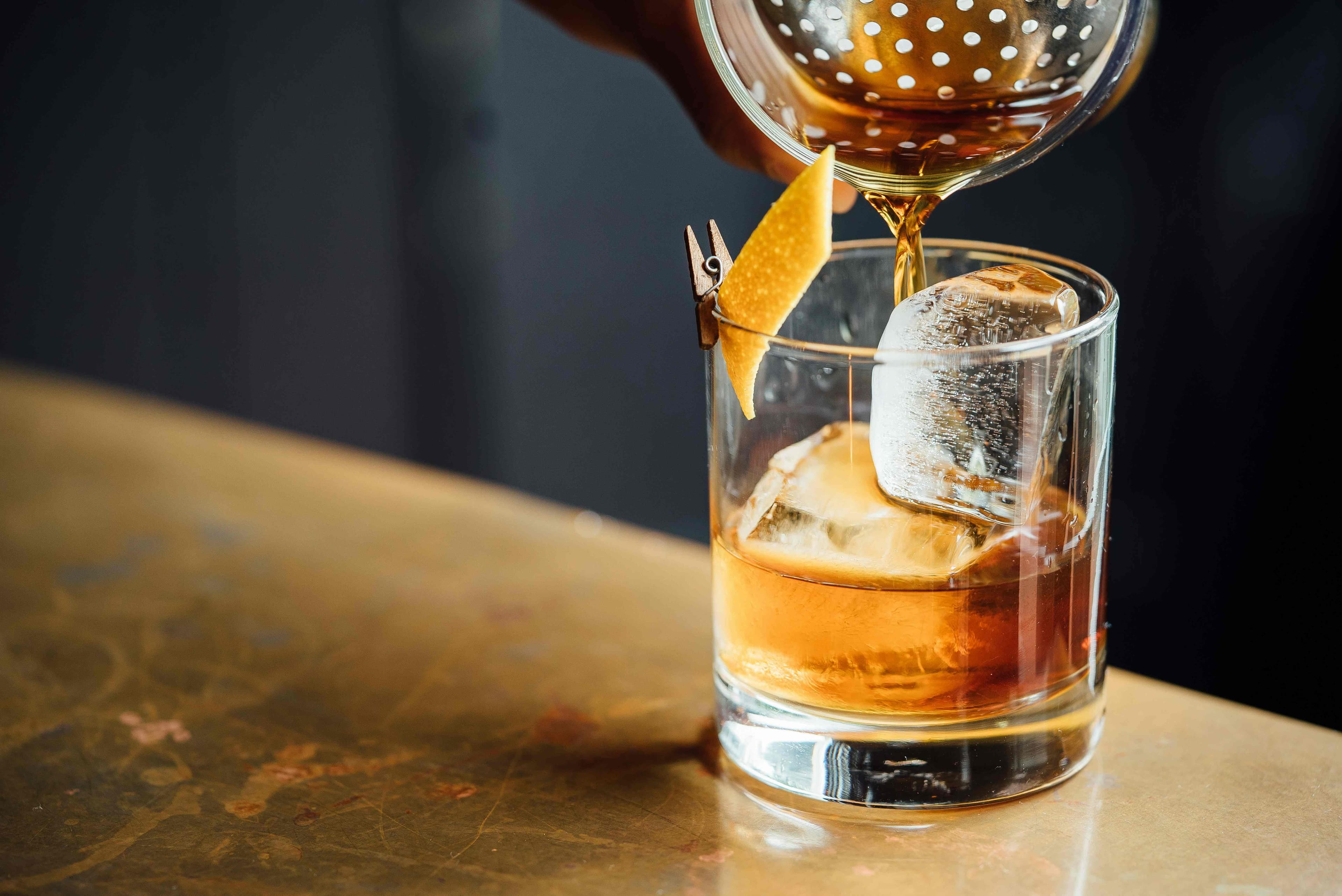 Cocktail ambrato servito in un bicchiere basso con grandi cubetti di ghiaccio, guarnito con scorza d’arancia tenuta da una mini molletta, mentre viene versato con un colino da barista su un bancone dorato.